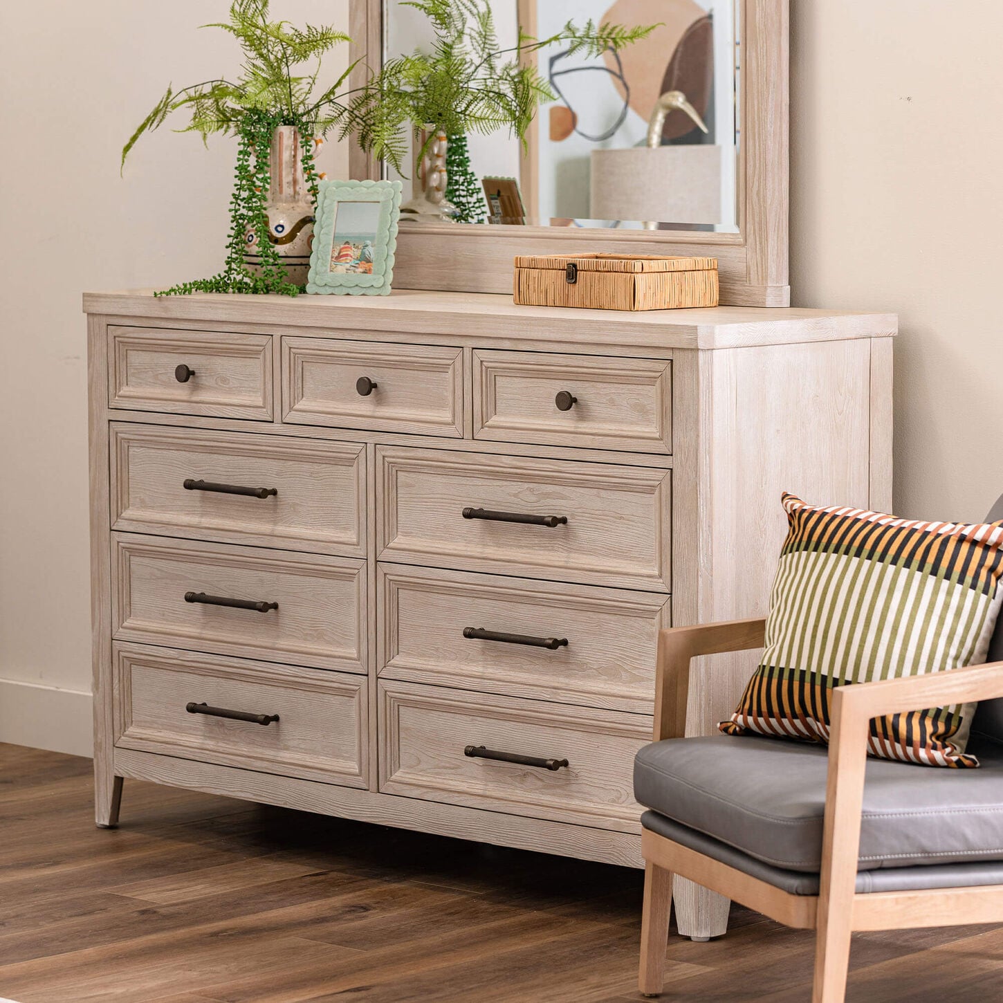 Light wood dresser with nine drawers, mirror, plants, and photo frame next to chair with striped pillow.