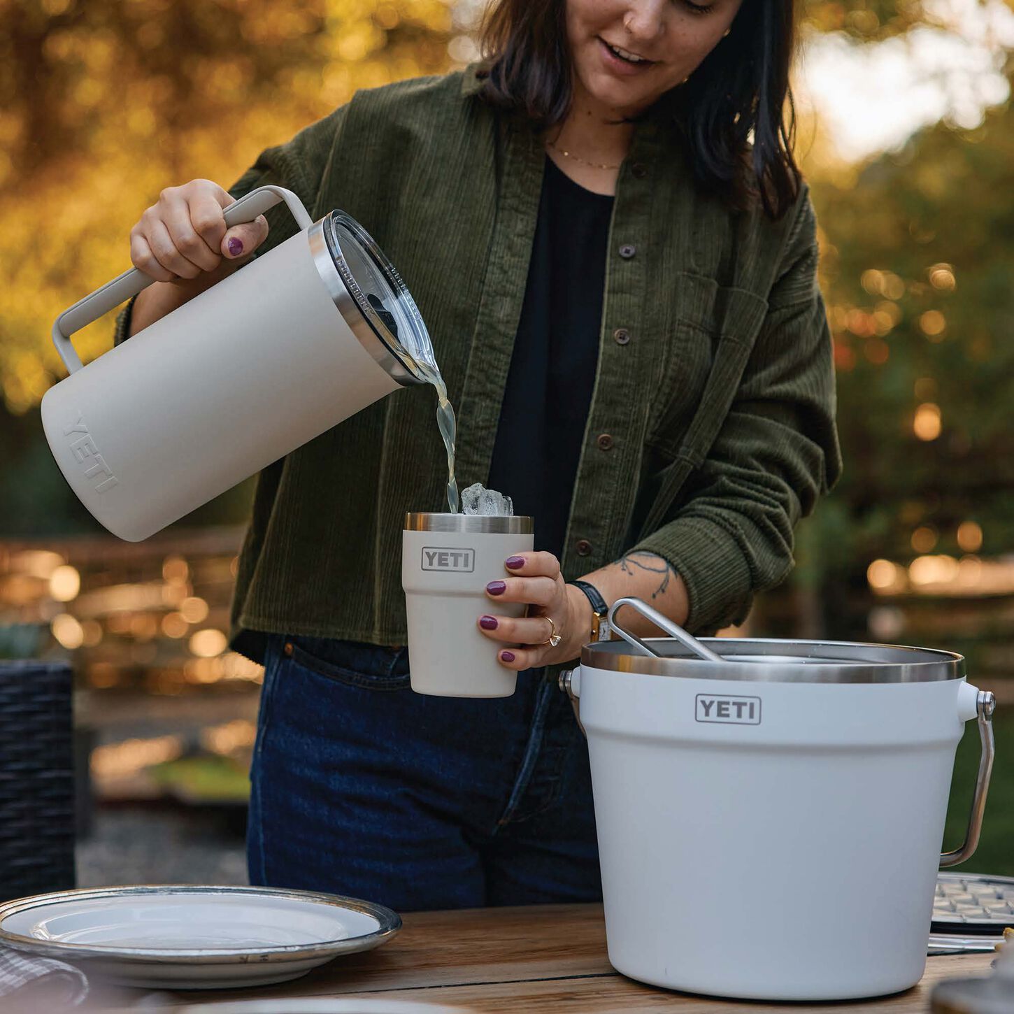 lady pouring liquid from a white yeti pitcher to a white yeti tumbler glass in front of a table with a plate and a white yeti bucket sitting on it