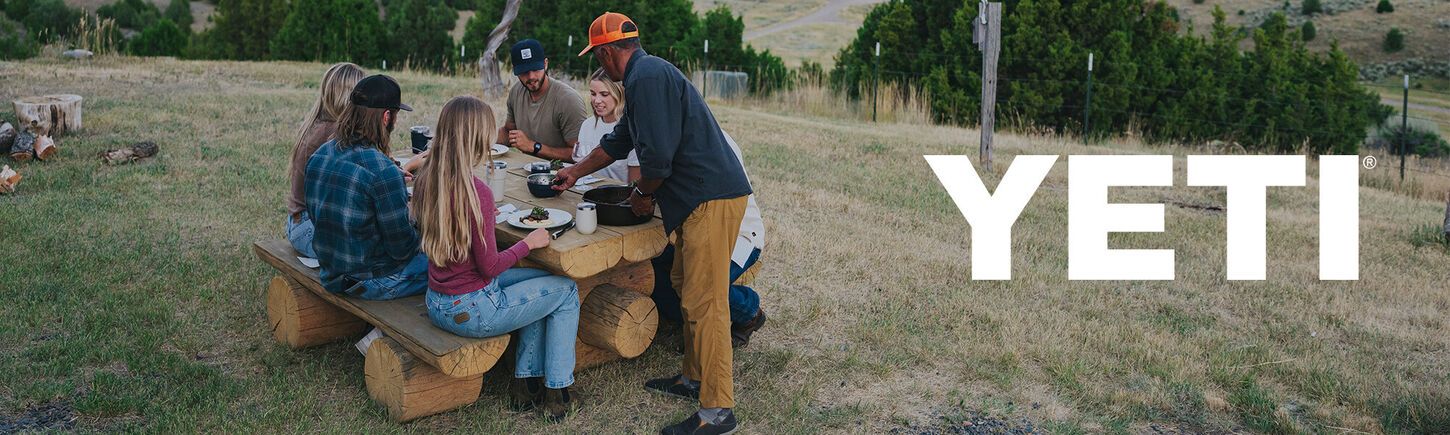 people gathered at a picnic table made from logs on a grassy hill and the work yeti over the top of the picture