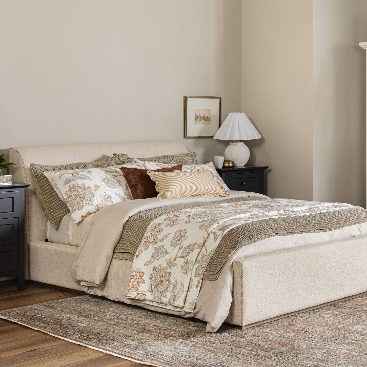 Neutral bedroom with upholstered bed, floral quilt, layered pillows, black nightstands, and area rug.