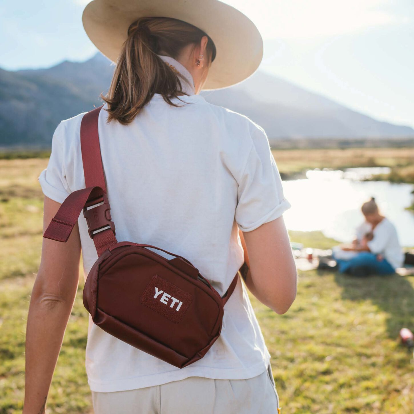 person wearing a white shirt carrying a maroon yeti sling bag walking away on the side of a stream