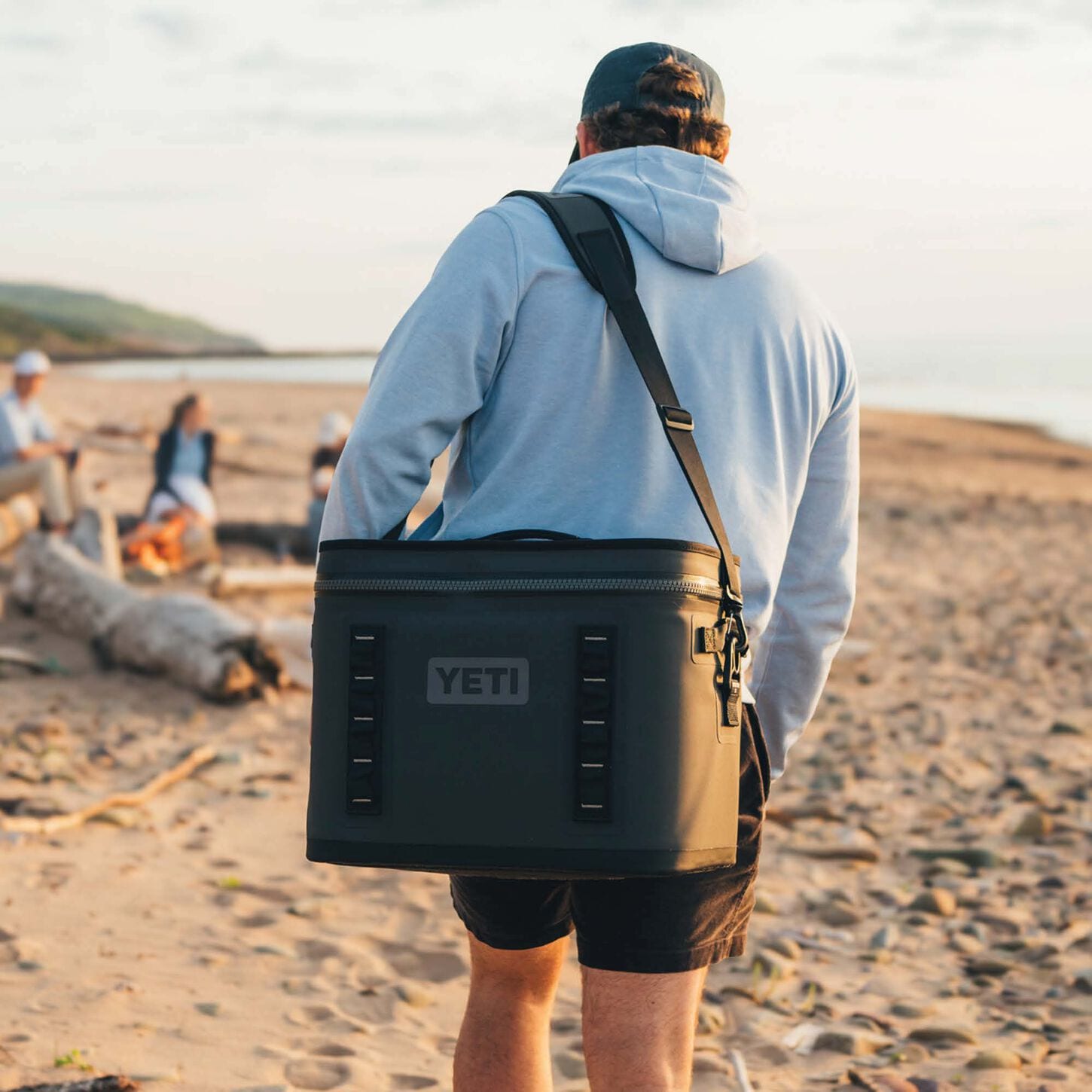 person in a blue hooded sweatshirt carrying a grey yeti cooler walking down the beach