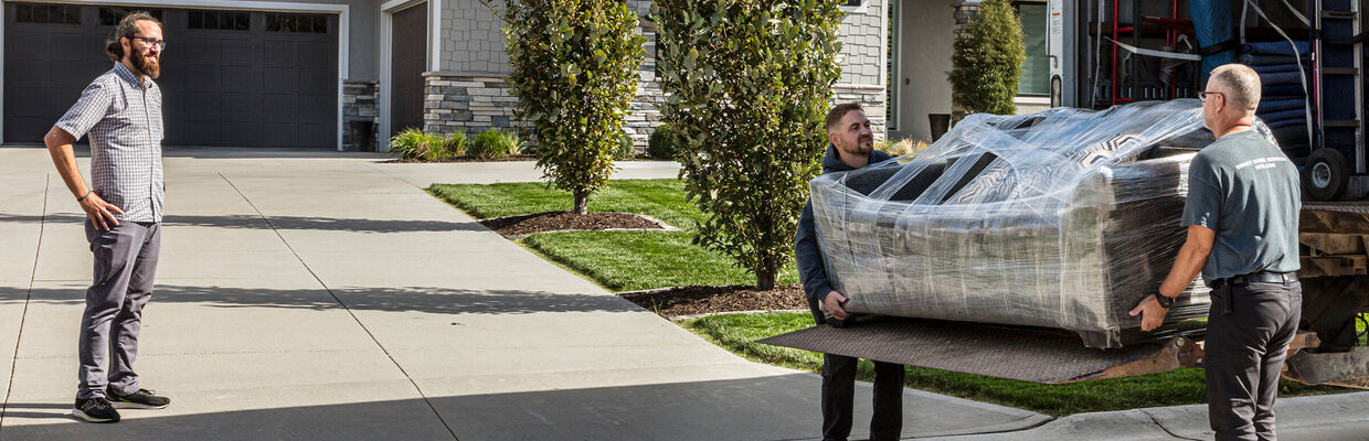 Delivery people unloading a sofa from the truck while a customer looks on in front of their home