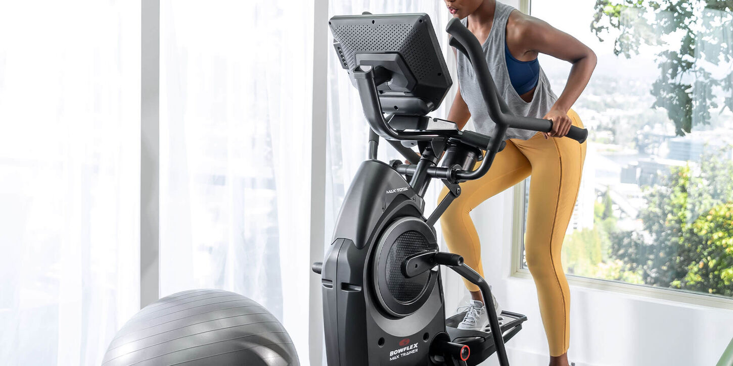 Someone on a elliptical in their home gym with an exercise ball and windows in the background.