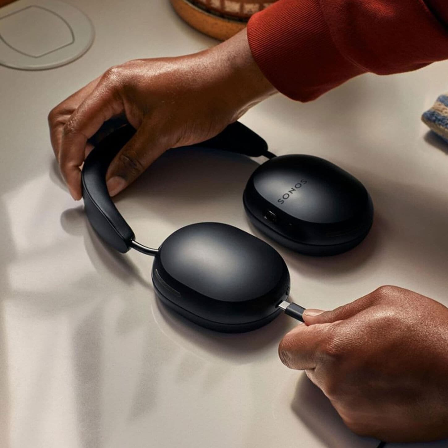 Hands holding black Sonos over-ear headphones on a light surface, with charging cable connected.