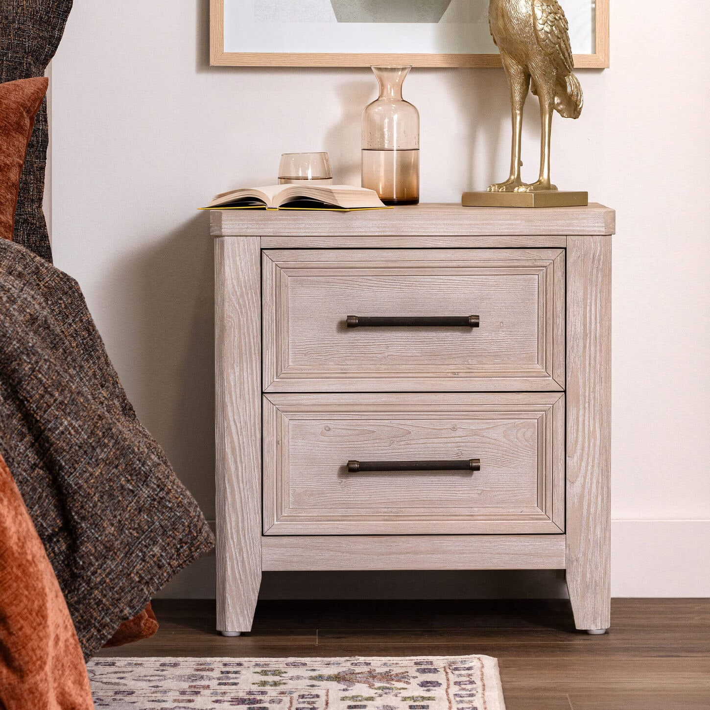 Light wood nightstand with two drawers, open book, glass vase, gold bird sculpture, and framed wall art.