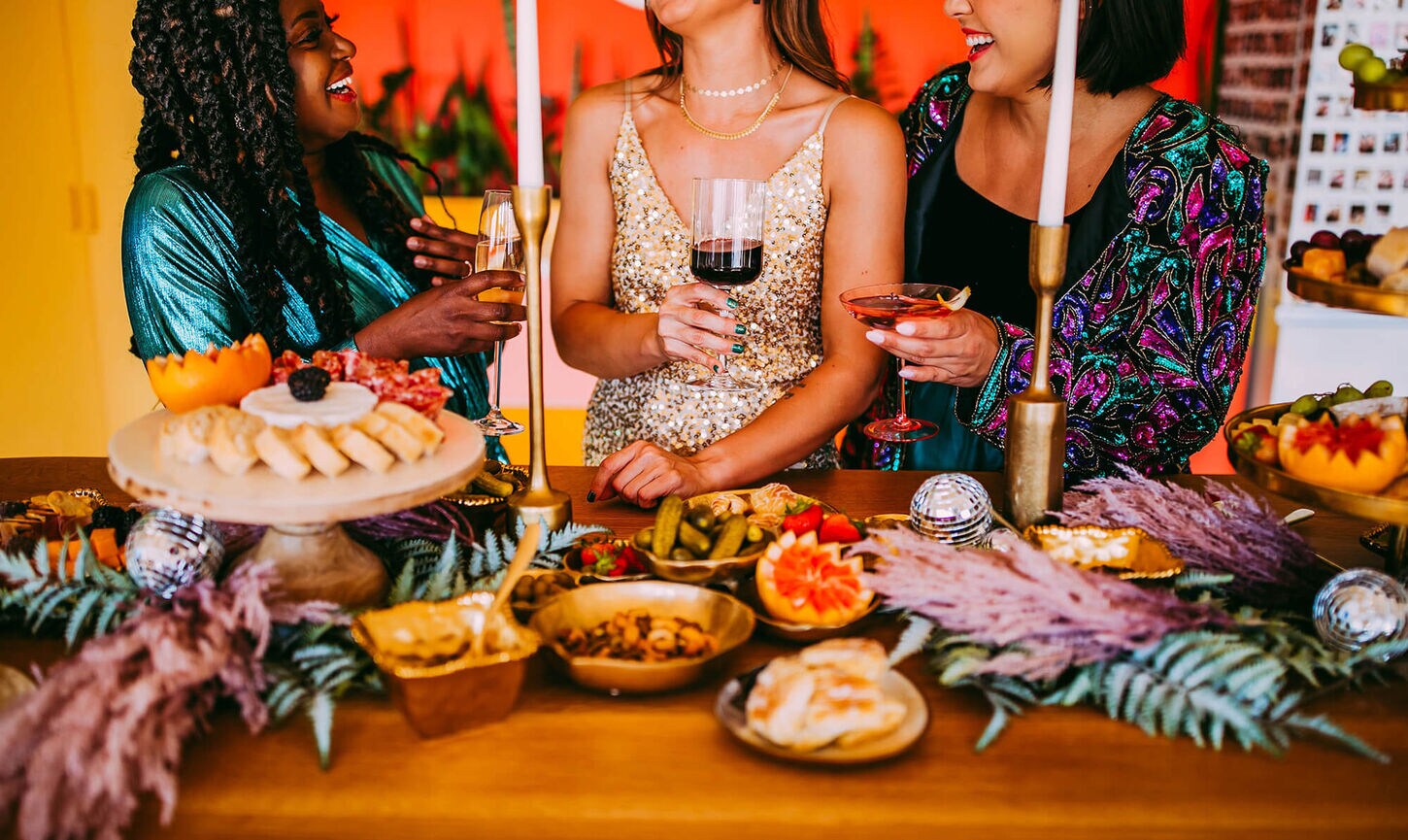 3 women gathered around a bar top with food, candle sticks, serve ware, and drinks.