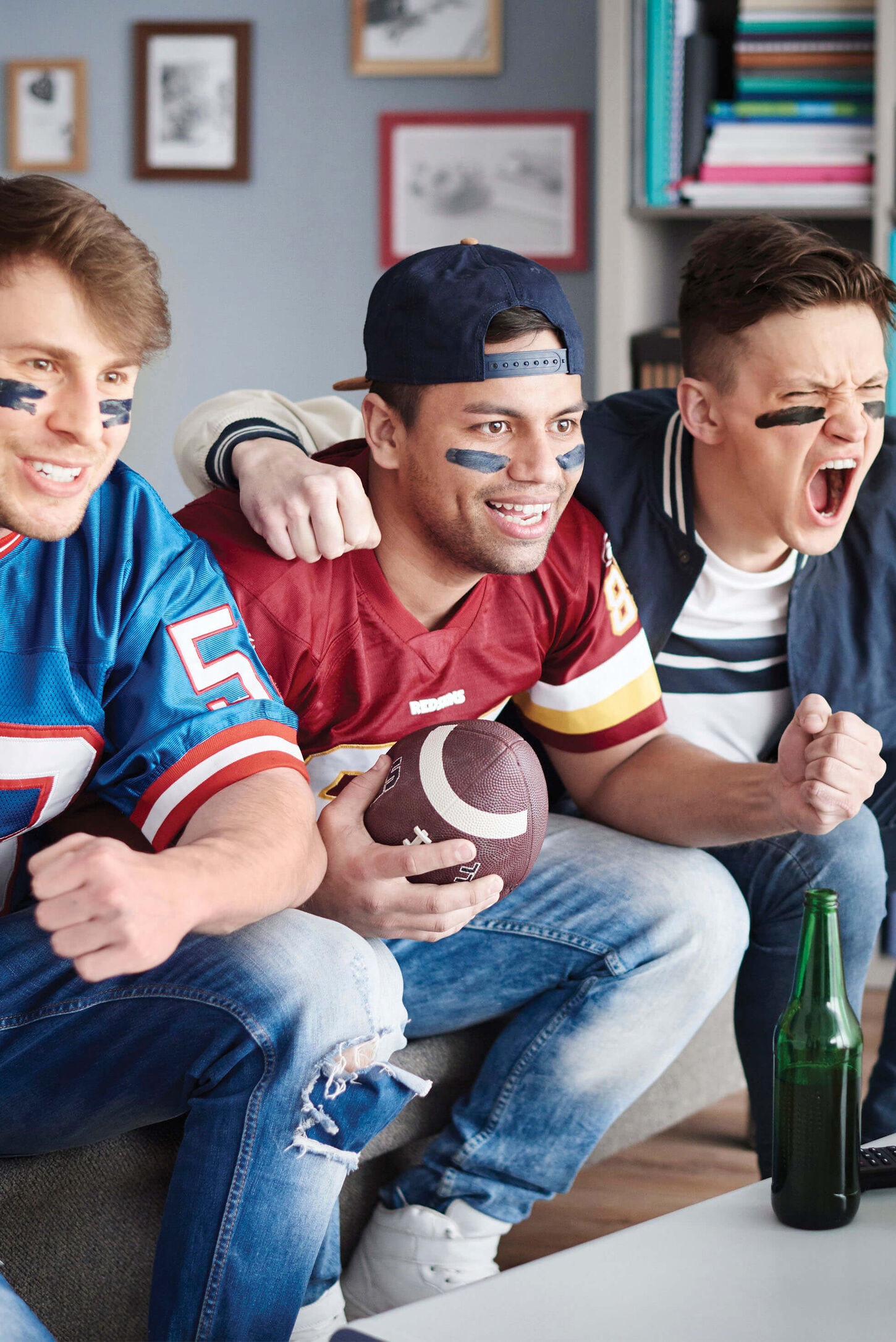 3 men sitting on a couch with a football in hand and watching a game on tv
