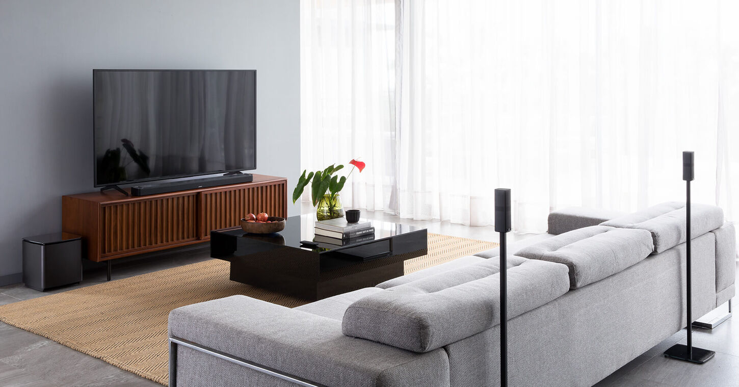 A grey walled living room with speakers behind the couch and a sound bar underneath the TV.