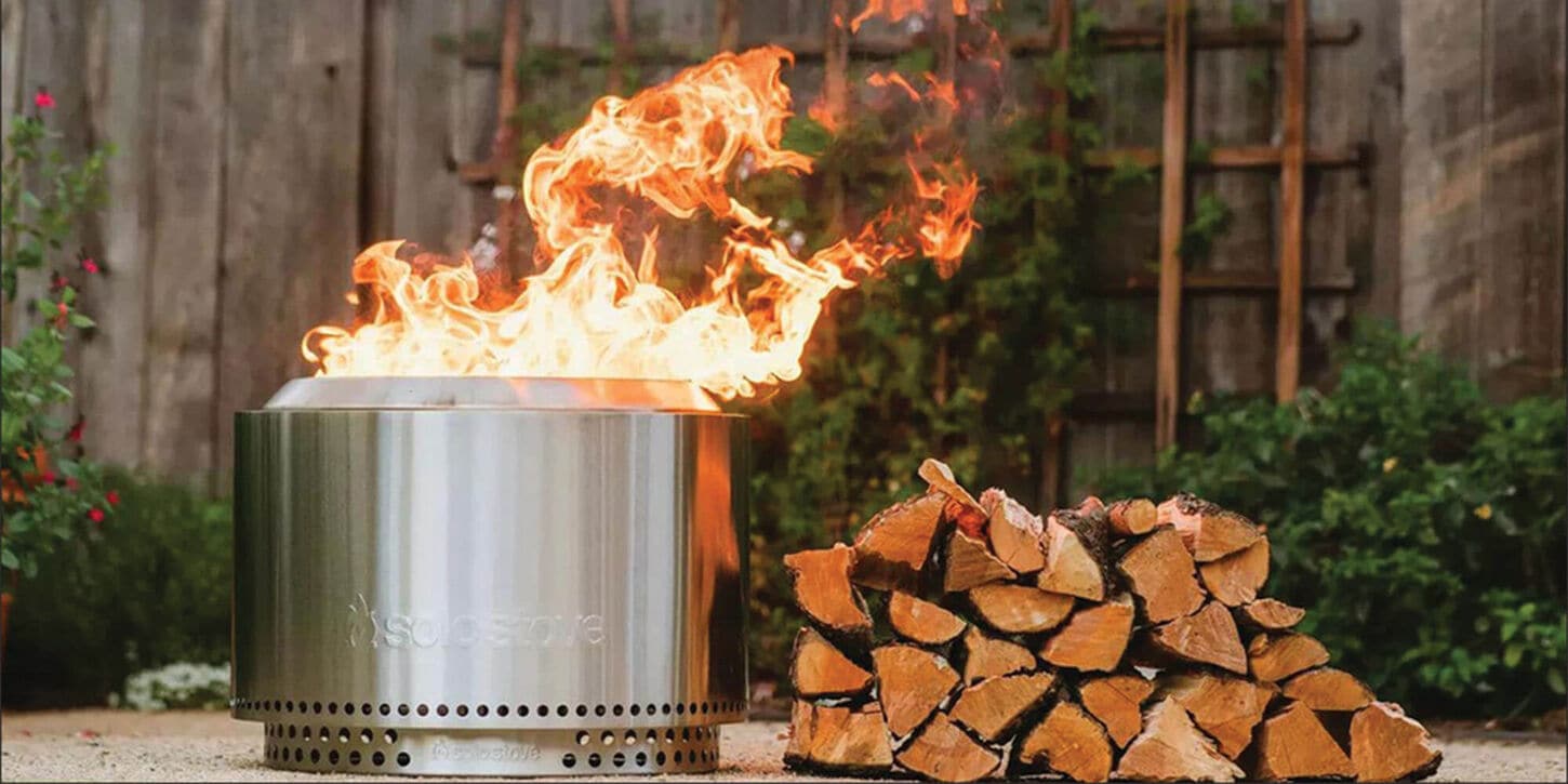 Solo Stove firepit in flames next to a pile of wood in the backyard
