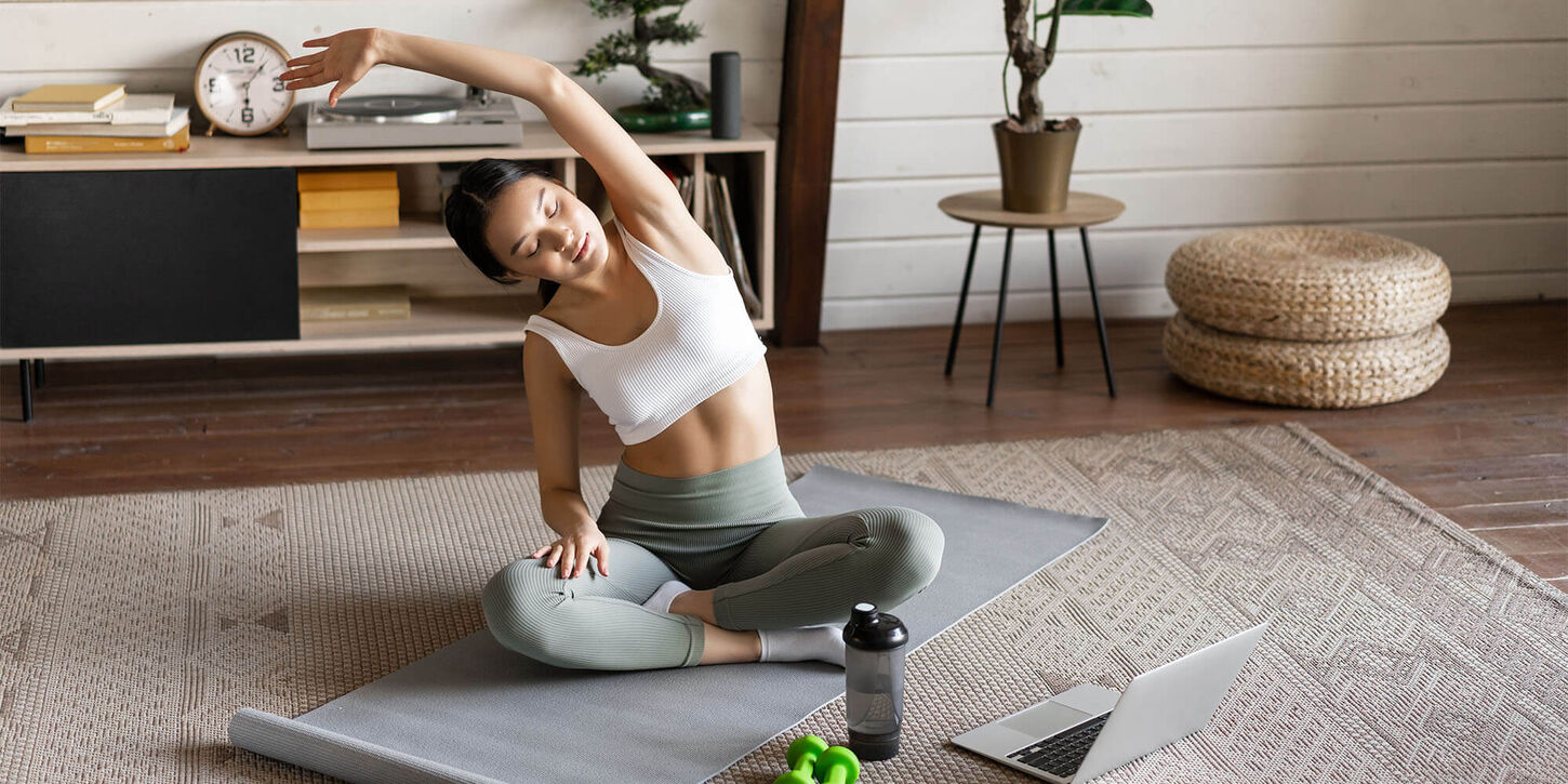 Girl sitting on the floor on a yoga mat in her home gym with workout equipment and furniture in the background.