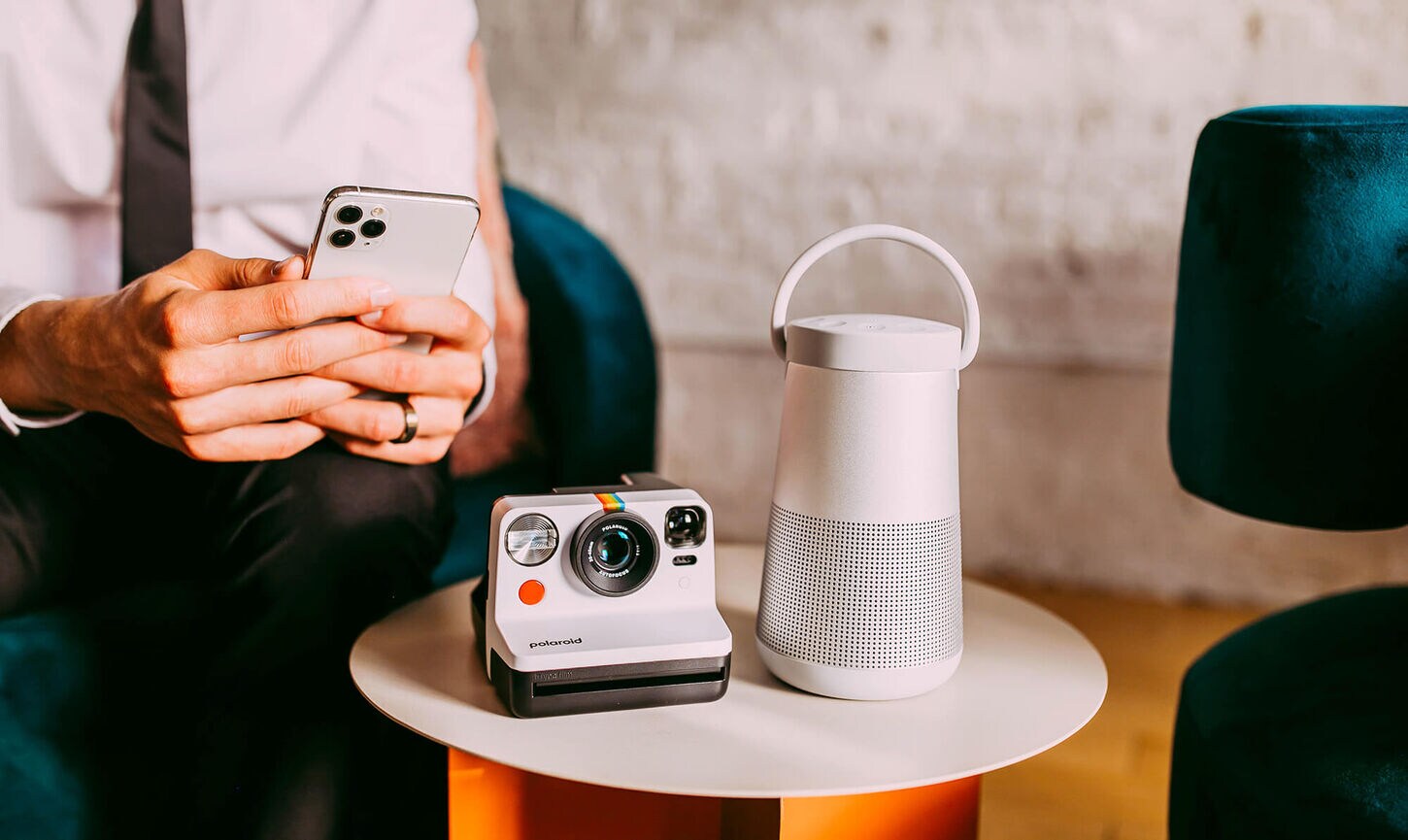 A man holding a phone trying to connect it to a Bluetooth speaker and right next to it is a polaroid camera.