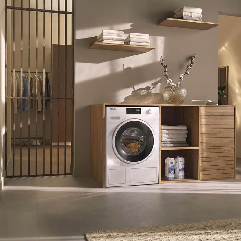 miele washing machine in white under a wooden cabinet with a shelf on the right and a door way to the left 